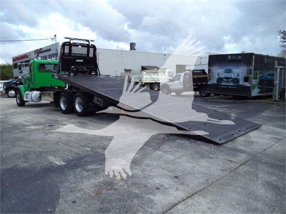 Flatbed tow truck with ramp extended, 2014 Peterbilt 367 logo visible on truck; green tow tractor in background; cloudy sky.