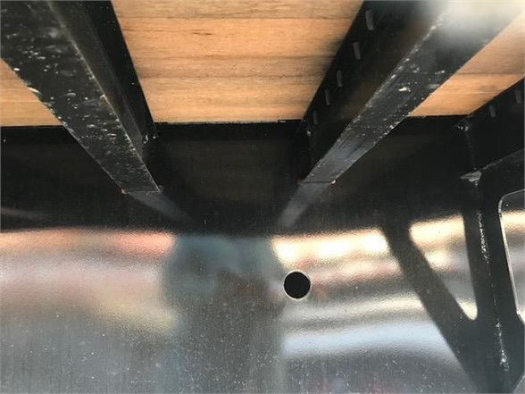 Flatbed trailer undercarriage with wooden deck and black steel beams; a circular hole visible in shiny metal surface.