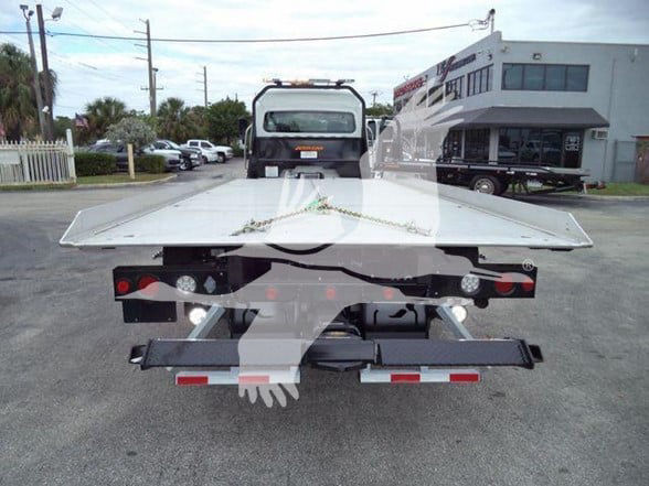 Freightliner M2 106 flatbed truck rear view with chrome platform, tow setup, and tail lights. Stock number JU6165FM visible on the back. A building and other trucks in background.