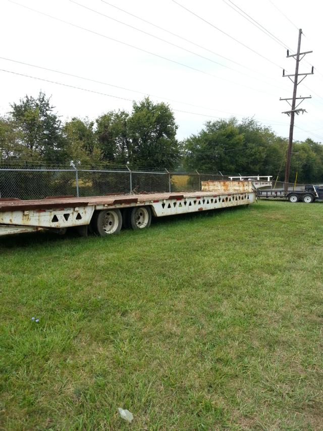 Lowboy trailer with multiple axles showing rusted deck and side rails, open field with fence in background.