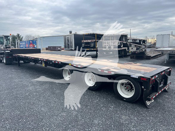 3-axle steel drop bed trailer in a yard, long flatbed with wood deck, dual rear axles and gooseneck area visible.