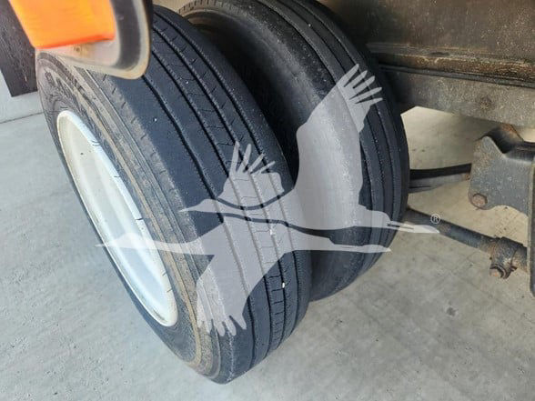 Trailer wheel with dual tires and hub, part of flatbed trailer. Visible details include tandem axle, leaf springs, and frame near the wheel.