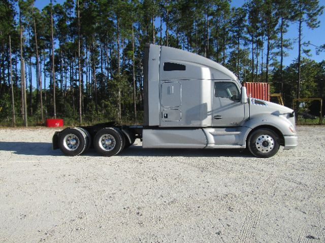 Side view of a silver 2020 Kenworth T680 tractor in a gravel lot with trees in the background.