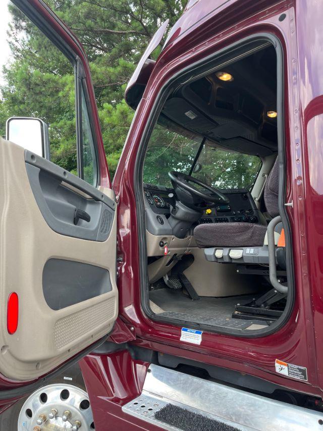 Truck cab interior of 2015 Freightliner Cascadia showing driver seat, steering wheel, dashboard, and open door with beige interior panels.