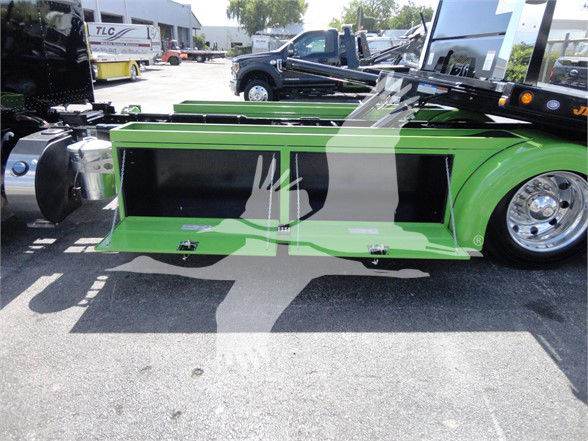 Green Peterbilt 337 truck side view showing aerodynamic side skirts with black panels and chrome wheels, front wheel, and surrounding lot with other trucks.