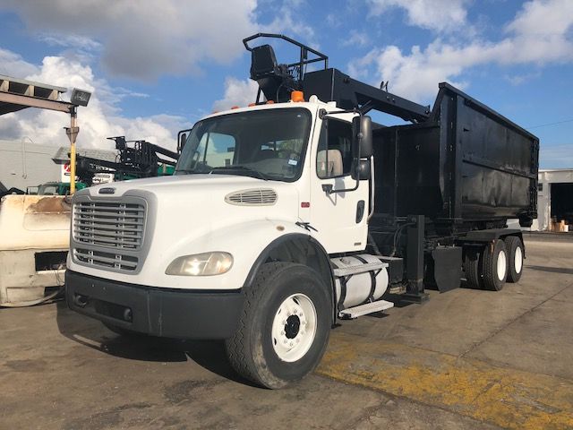 Grapple truck: white Freightliner M2-112 with black dumping bed, front view showing cab, grille, and side steps.