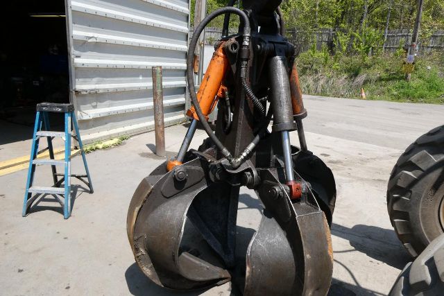 Excavator clamshell bucket attachment with hydraulic lines and steel jaws near a shipping container.