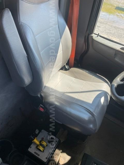 Truck cabin seat and pedal area of a 2012 Peterbilt 365 showing a gray driver's seat and steering wheel with floor mat and control box near the seat.