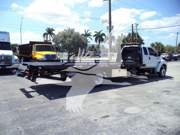 Ford F650 tow truck with flatbed trailer hitched to white pickup truck, in a parking lot, palm trees in background.