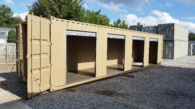 Large beige cargo container trailer section with four open bays on a gravel lot, blue sky and trees in background.