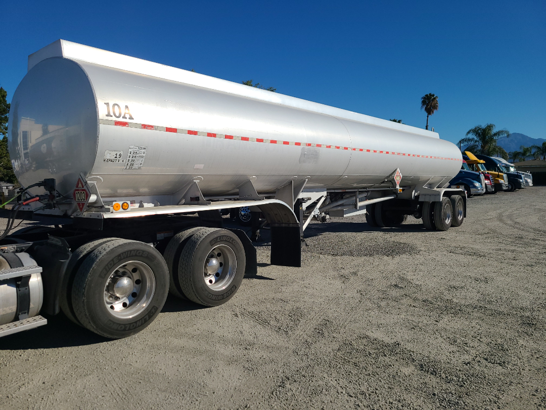 Semi-trailer tank with cylindrical aluminum tank on a multi-axle tractor, long shadow, red reflective stripe along side, 1203 hazard placard on rear, blue sky.