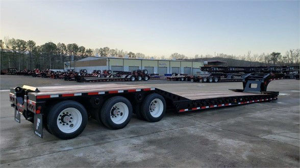 Fontaine lowboy trailer showing triple axle front half and long low deck with roller paver on the rear; wide flatbed with black coating and white rims.