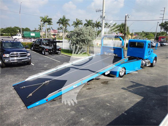 Blue Peterbilt tow truck with a long tilt bed in a parking lot.