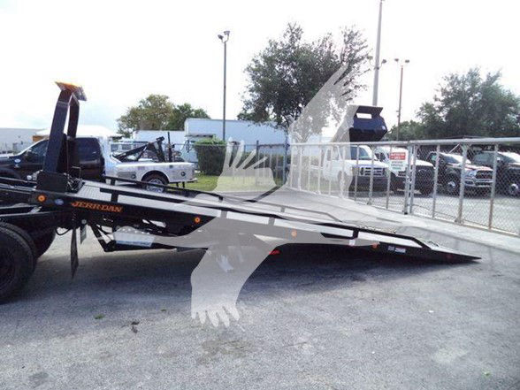 Tow truck flatbed trailer with black and orange branding showing a long hydraulic tilt bed and ramps in an outdoor lot.