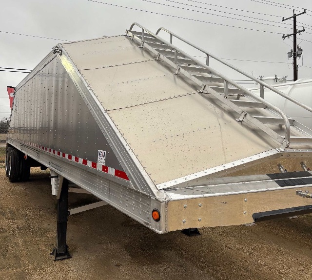 Aluminum hopper trailer with a raised grain chute, side ladder and reflective tape along the lower edge. Visible bolts and rivets on the aluminum panels.