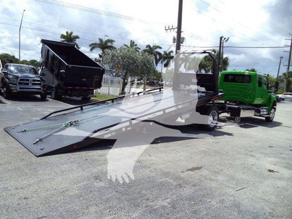 Truck tow truck with a hydraulic car carrier ramp extended, International MV stance, green truck in background.