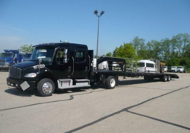 Freightliner M2 heavy-duty truck with a flatbed trailer carrying a car, black cab, daytime running lights, yard in background.