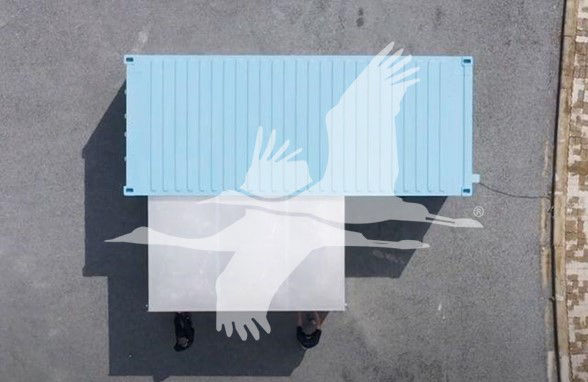 Aerial view of a light blue rectangular trailer with a white ramp or extension, casting shadows on a gray surface; no legible labels visible.