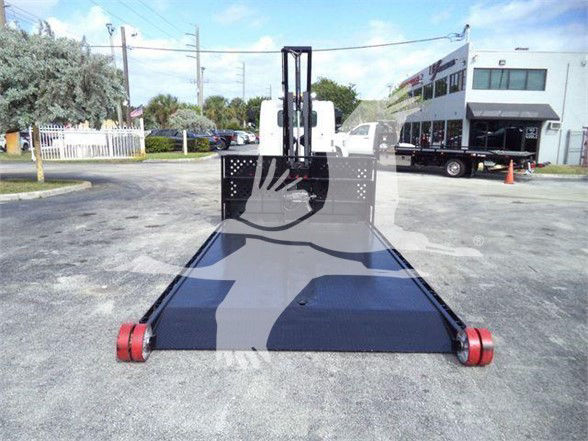 Flatbed tow truck pallet with hydraulic lift and fork attachment in front, black platform with red rollers, truck yard in background.