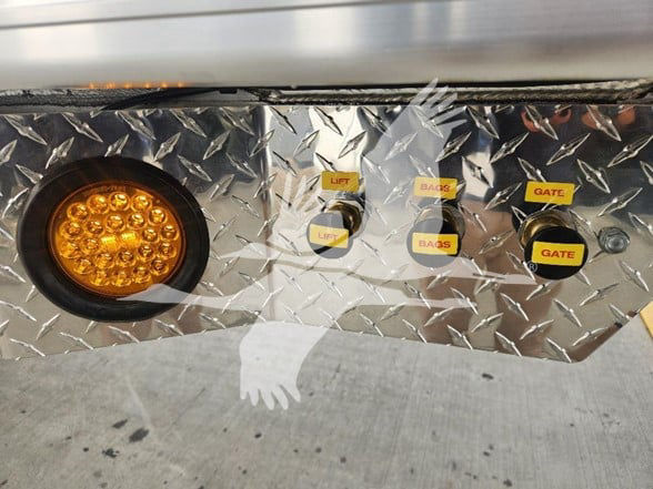 Trailer side panel showing control switches with yellow labels: LIFT, BIAS, GATE. Round amber LED work light at left. Diamond plate aluminum surface.