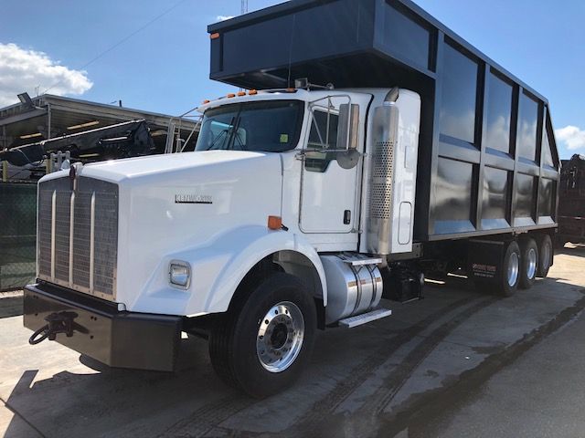 Kenworth T800 grapple dump truck in white with a large black dump bed, front grille, chrome exhaust stacks, and six wheels visible.