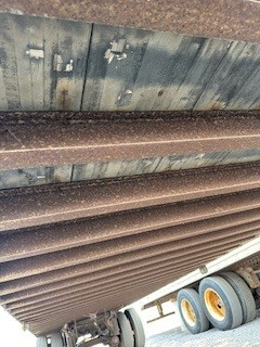 Dented corrugated metal trailer wall with support beams and wheel assembly visible, showing rust-colored metal surface and stacked sections.