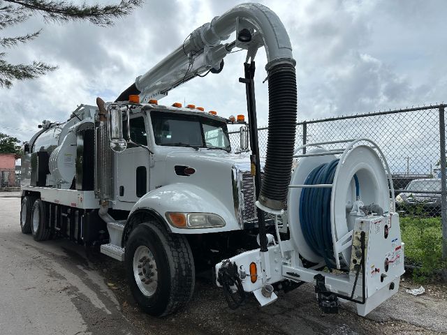 Peterbilt vacuum truck showing large hose reel on rear with extended boom and blue hose. Front cab is white with chrome grille and multiple amber clearance lights.