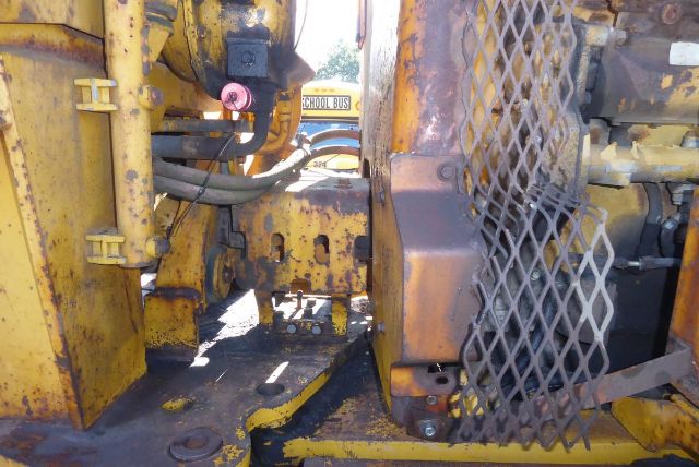 Caterpillar 16G machine bay showing yellow metal framework and a mesh guard with hydraulic lines and rust-covered surfaces.