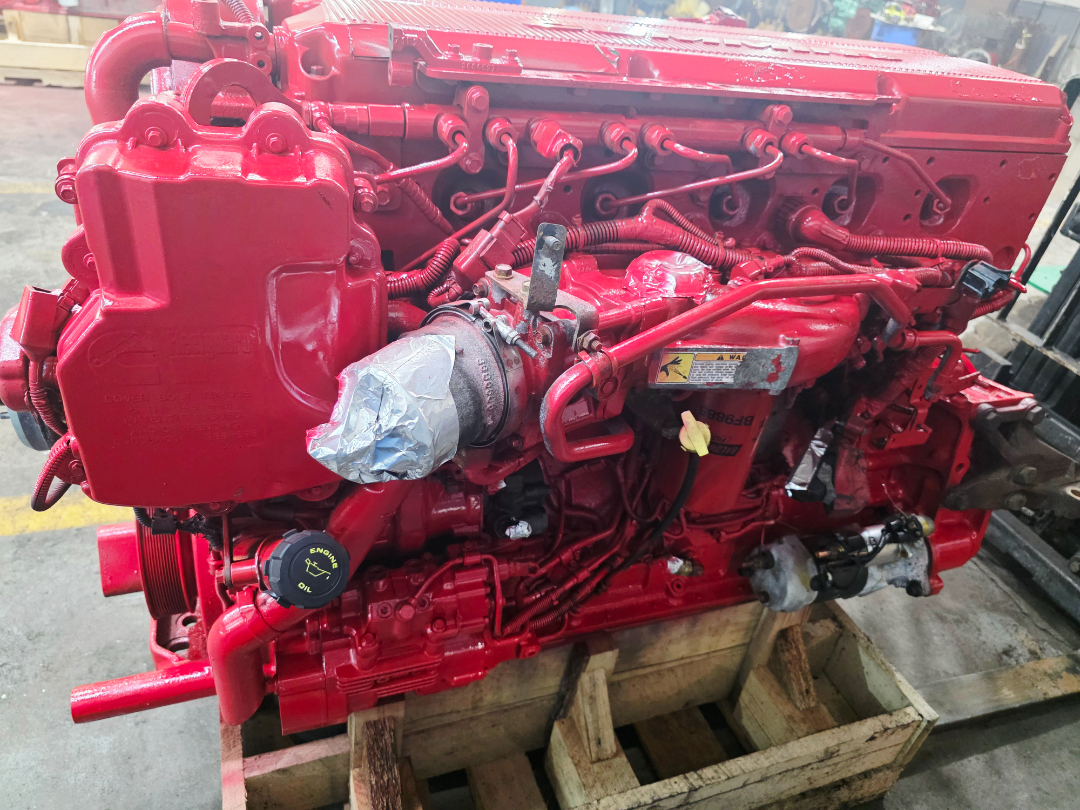 2014 ISX15 engine with red paint showing intake manifold, turbocharger, and various hoses; label stickers on side, oil cap visible.