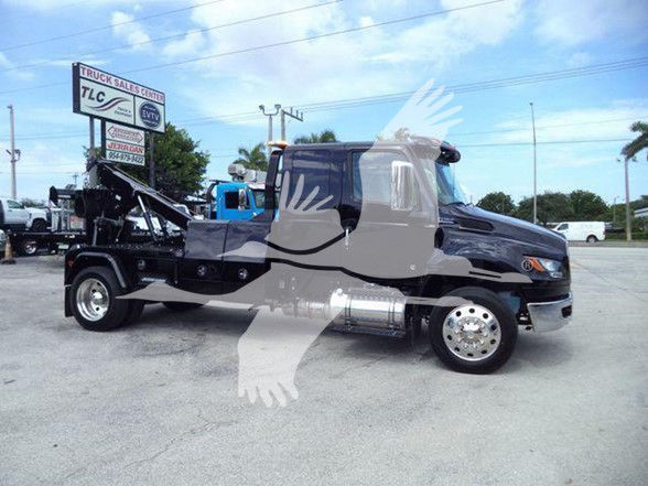 Truck bay showing 2025 International MV Tow truck with chrome fuel tanks, black hood, gray cab, and spare parts on the flatbed area. Visible text on a sign: TLC Truck Sales Center.