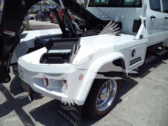 Truck bed with operator controls and tow equipment on a white 2023 International CV tow truck, partial rear wheel visible, black hydraulic arms and control panel in foreground.