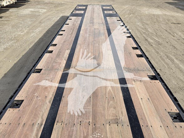 Tri-axle drop deck trailer bed with wooden planks and black edge rails, viewed from end, long perspective.