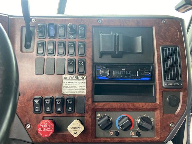 Truck dashboard panel with multiple switches, wood-grain finish, JVC stereo, climate controls, warning label, and parking brake knob.