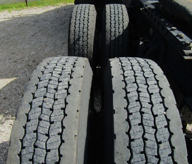 Kenworth T680 cab and chassis wheelset showing four large rear dual tires with tread pattern, roadside gravel, and chassis frame visible.