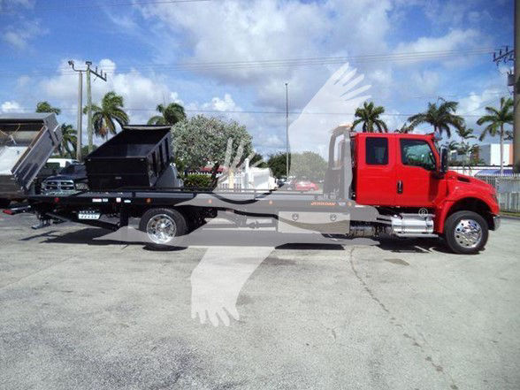 Tow truck with red cab and long flatbed, loading a black dumpster in a parking lot, palm trees and blue sky in background.