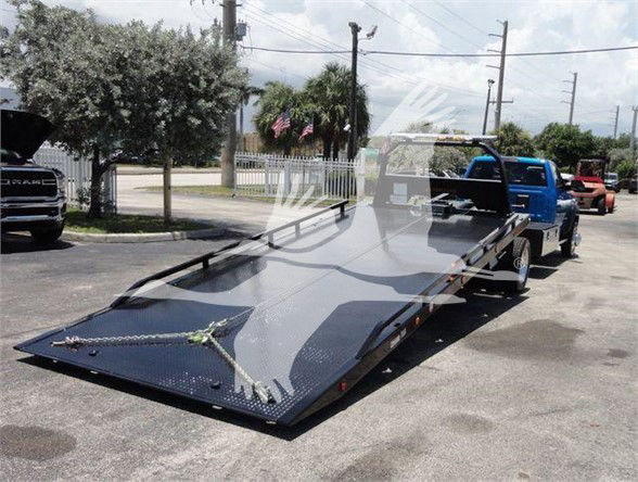 Tow truck flatbed trailer being towed by a blue Ram pickup, black metal platform with ramp at front, street scene in background.
