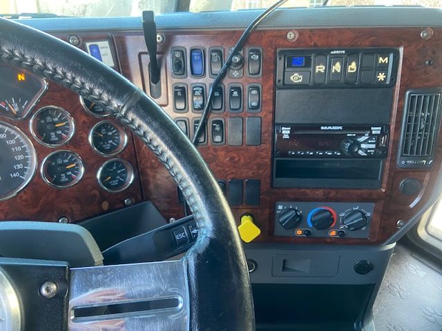 Dashboard of a 2015 Mack CXU613 showing instrument cluster, wood-grain panel, and controls including RN S shift indicator, climate controls, and a stereo unit. Visible text includes 'MACK' on the radio and various labeled buttons.