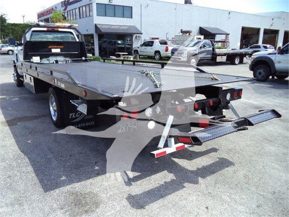 Tow truck flatbed trailer with ramp in rear and SUV/ trucks in background.