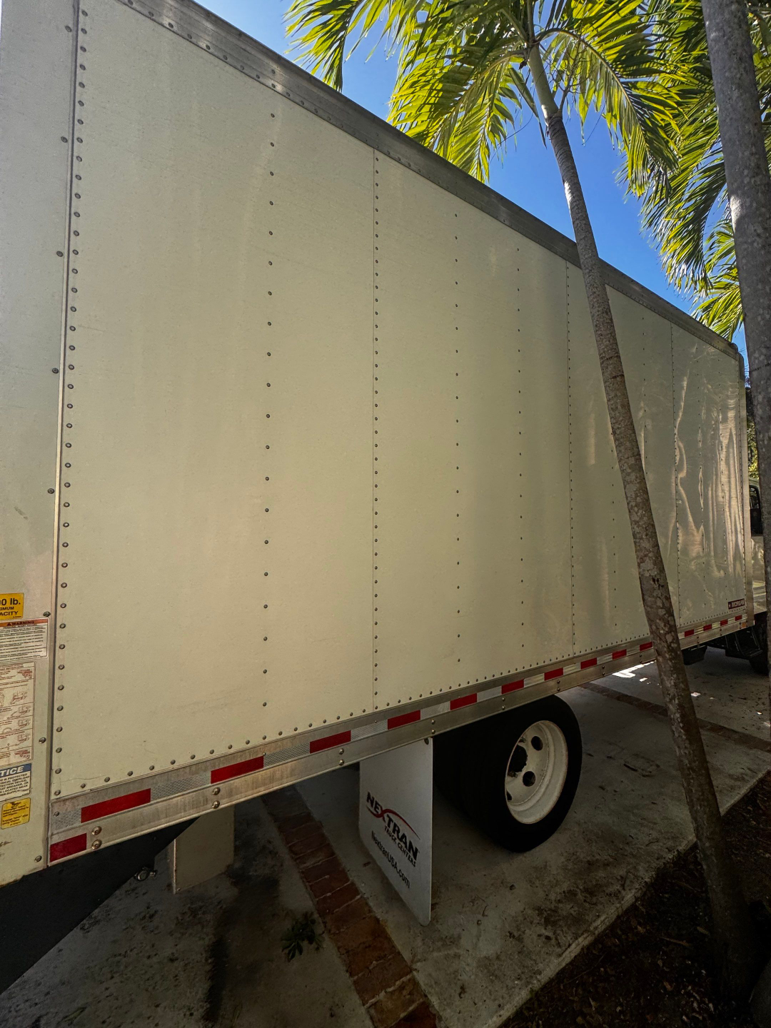 Box truck trailer with smooth white panels and riveted seams, Isuzu NQR in 2022. Side view shows wheels, red/white reflective striping, and a palm tree shadow.