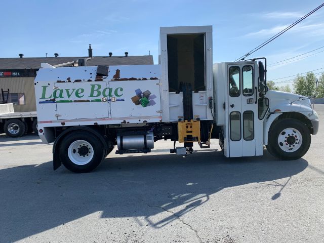 Truck side view of a white 2006 International 4300 garbage truck with a Lave Bac recycling body. Side door open, graphic 'Lave Bac' on the side, gas tank and machinery under the body visible.