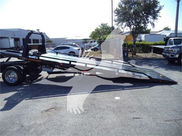 Tow truck flatbed trailer attached to a Ram 5500 SLT, side view showing extended steel ramp/haul bed with wheel chocks and parking lot backdrop.