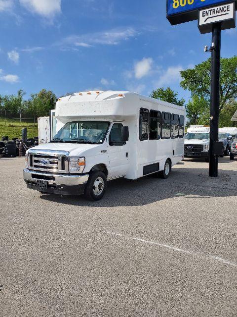 White 2025 Ford E450 shuttle bus in a dealership lot, front and side view, with blue sky and sign visible.