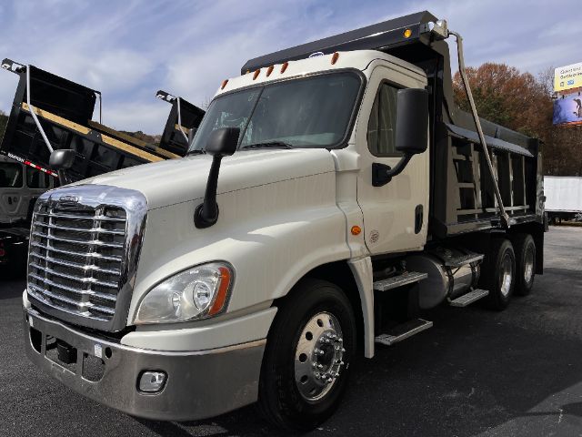 Freightliner Cascadia 125 dump truck in a light color, with a chrome front grille, large side mirrors, and a dump bed. Visible features include dual rear axles, fuel tanks under the cab, and a stepped side entry.