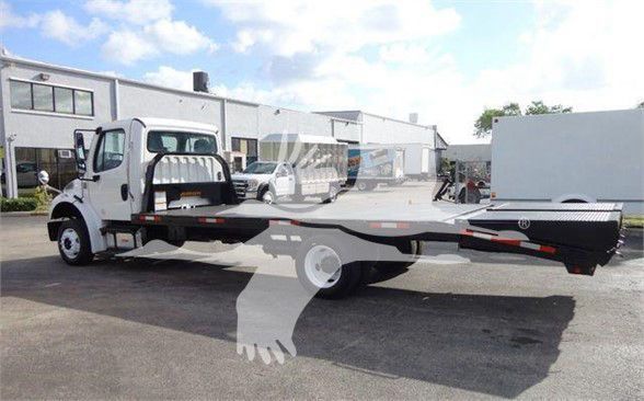 Freightliner Business Class M2 106 flatbed tow truck seen from side, white cab, long flatbed with wheel chocks and equipment storage on the back, truck in a dealership lot.