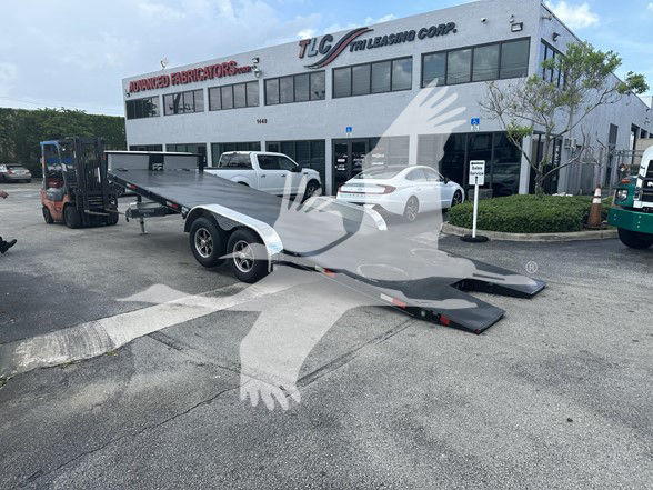 Trailer flatbed with lowered ramps in a dealership lot, TLC Trucking building in background.