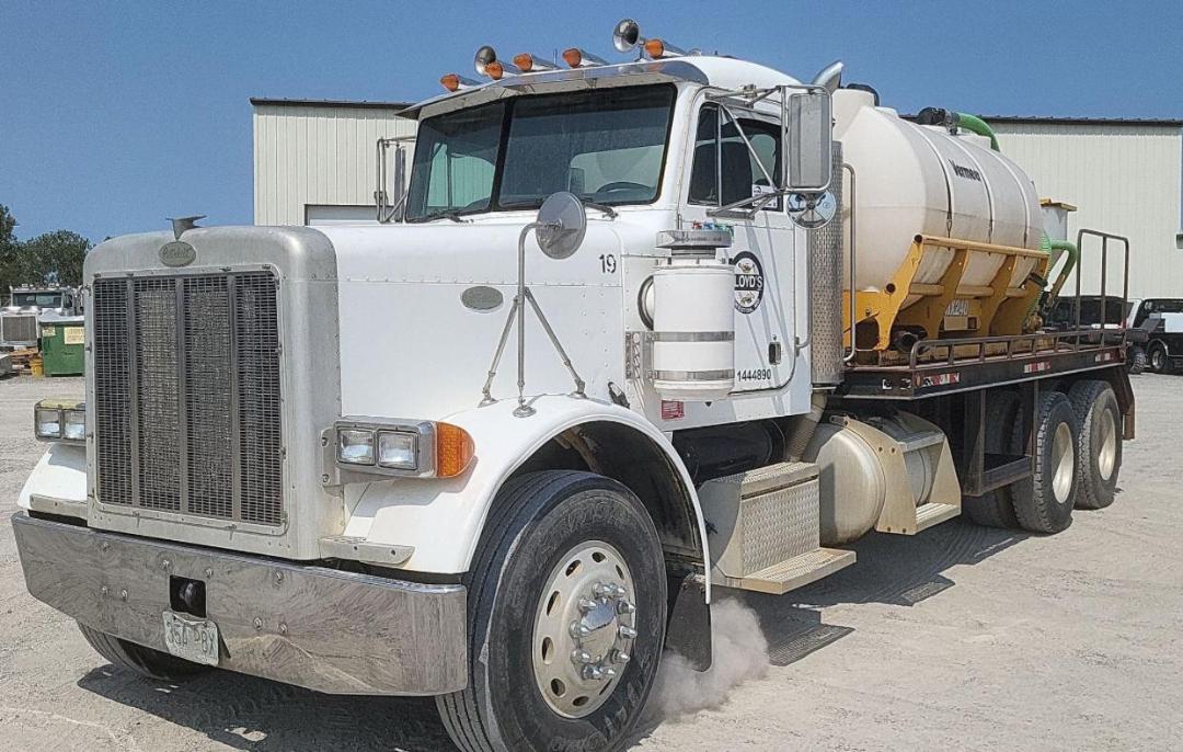 Peterbilt 379 heavy-duty truck with a white cab and a large chrome grille, air cleaners on the sides, multiple amber marker lights, and a tan/payload tank on a flatbed with dual-rear axles.