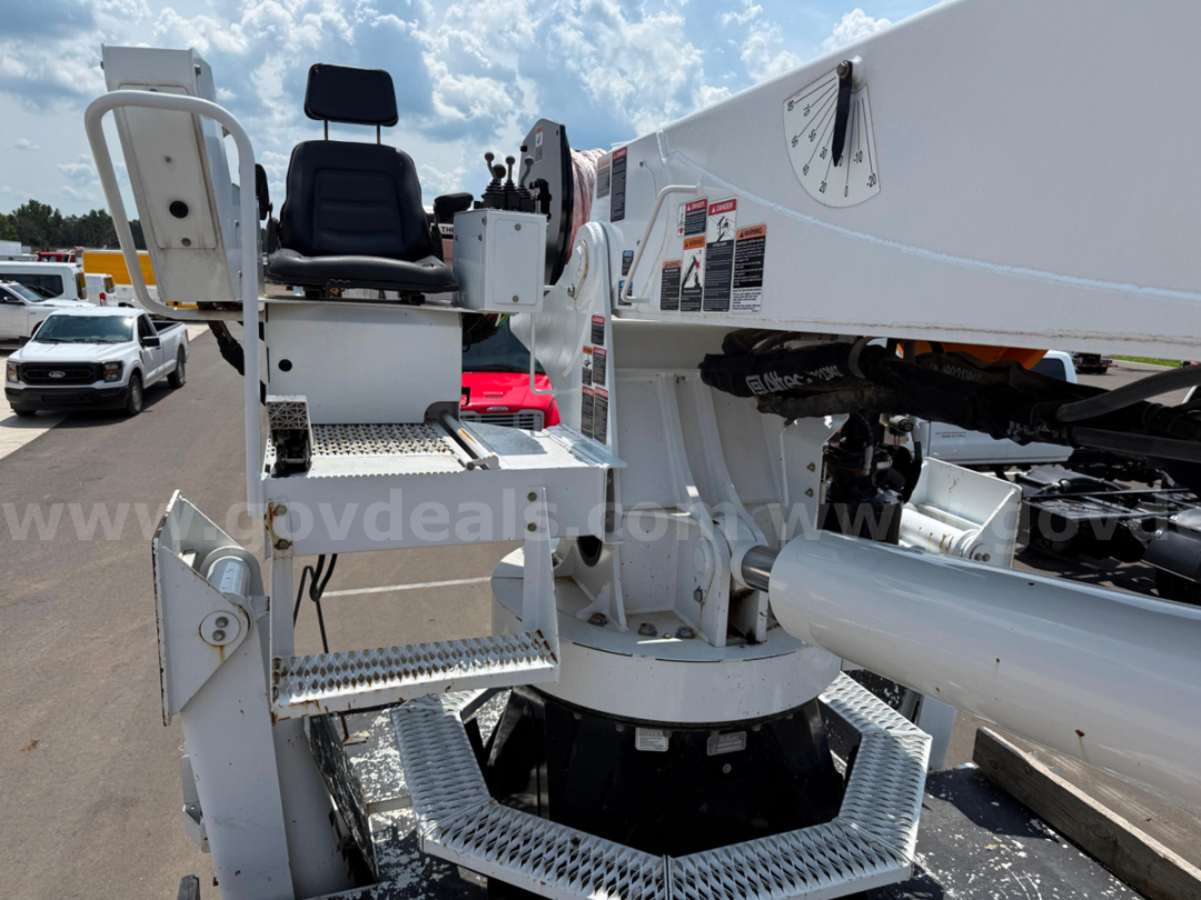 Truck-mounted aerial lift control platform with operator seat and joystick controls on a white boom arm. Platform includes step ladder and safety rails; hydraulic pivot and base visible.