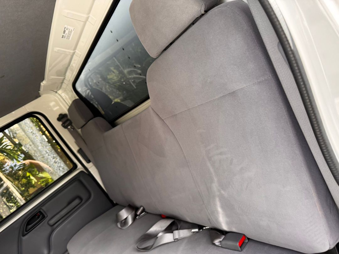 Isuzu NQR interior showing gray fabric bench seat with seat belts and side door panel. Passenger-side view with window and headrest. 