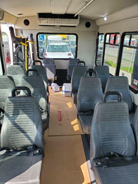 Bus interior with rows of gray seats, cardboard boxes on floor, rear air conditioning unit, windows on both sides.