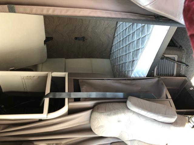 Truck sleeper cab interior showing bunk bed with quilted mattress, storage compartments, and passenger seat; beige upholstery and curtains.